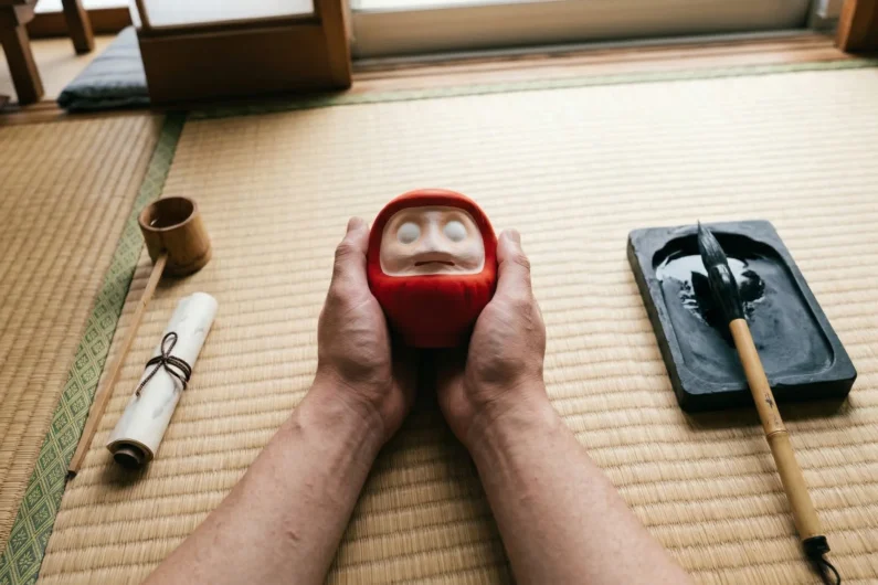 El mágico ritual del daruma y su significado real - como pintar ojo daruma metas Fotografía en primera persona de unas manos sosteniendo un daruma sin ojos pintados junto a un pincel de caligrafía y tinta negra sobre un tatami.