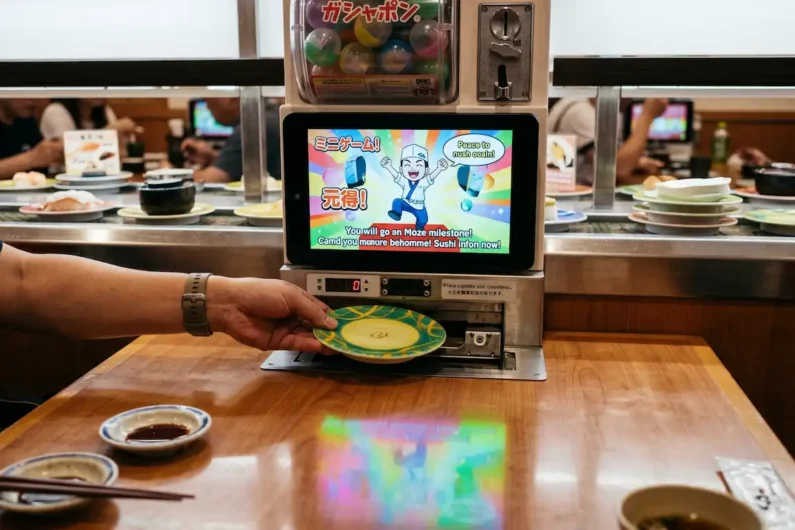 Guía de kaitenzushi (sushi de cinta) y trucos para aprovechar el buffet - bikkura pon kura sushi gashapon Mano insertando un plato vacío en una ranura de mesa de restaurante con una pantalla digital de premios.