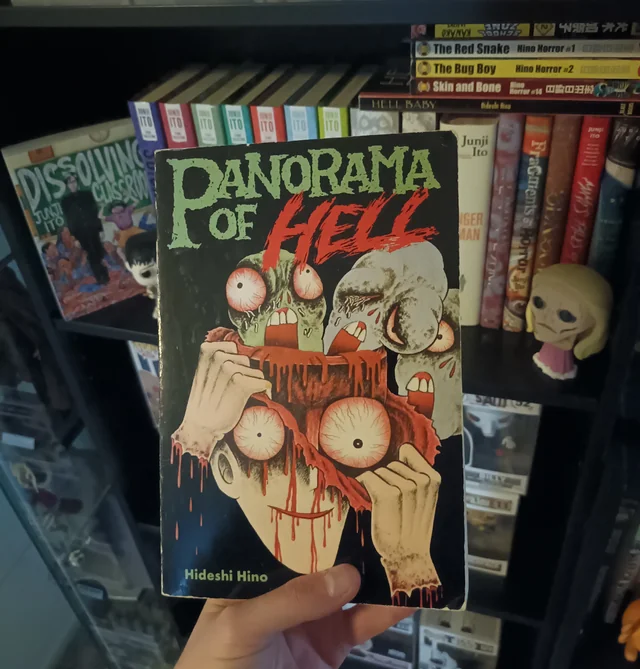 5 autores de manga de terror más perturbadores que junji ito - hideshi hino panorama of hell lectura fisica Fotografía de una mano en primera persona sosteniendo el tomo del manga panorama of hell de hideshi hino frente a una estantería llena de cómics.