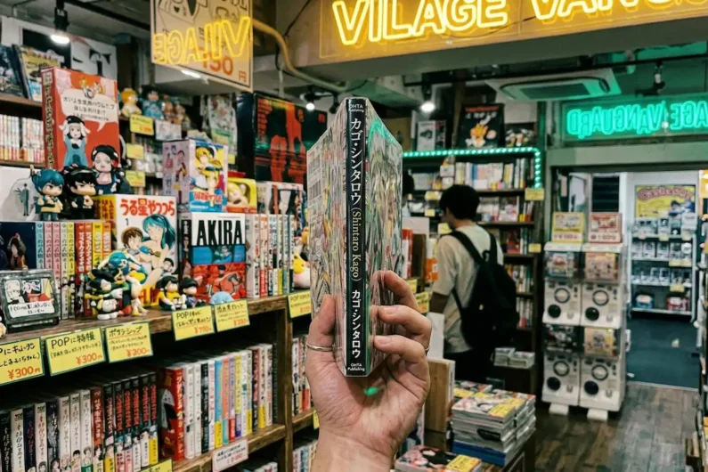 5 autores de manga de terror más perturbadores que junji ito - manga shintaro kago village vanguard shimokitazawa Una mano humana sosteniendo un grueso libro manga con portada brillante en una tienda oscura iluminada con neones de colores.