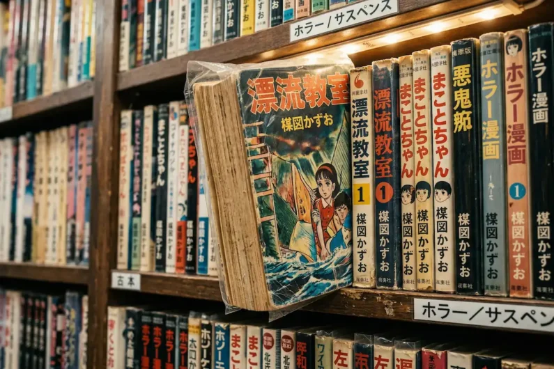 5 autores de manga de terror más perturbadores que junji ito - tomo original aula a la deriva kazuo umezu nakano Fotografía de un cómic manga antiguo envuelto en plástico arrugado descansando sobre una estantería de madera abarrotada.