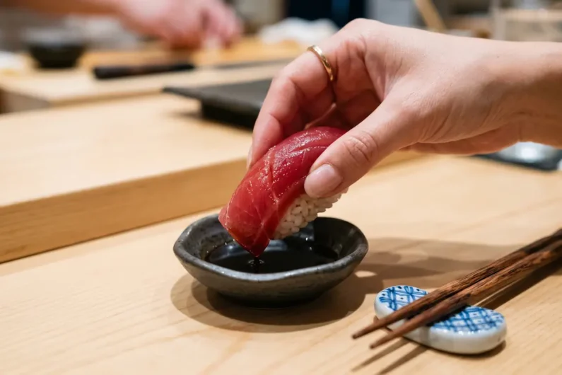 Los 10 errores más comunes al comer sushi en japón - como mojar sushi salsa soja correcto Una mano sosteniendo con cuidado un nigiri de atún de lado, tocando la salsa de soja exclusivamente con el pescado.