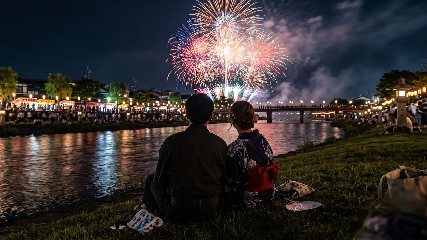 Vista trasera de una pareja vestida con yukatas tradicionales sentados en la hierba a la orilla de un río en japón, observando un gran espectáculo de fuegos artificiales multicolores sobre un puente iluminado con farolillos por la noche