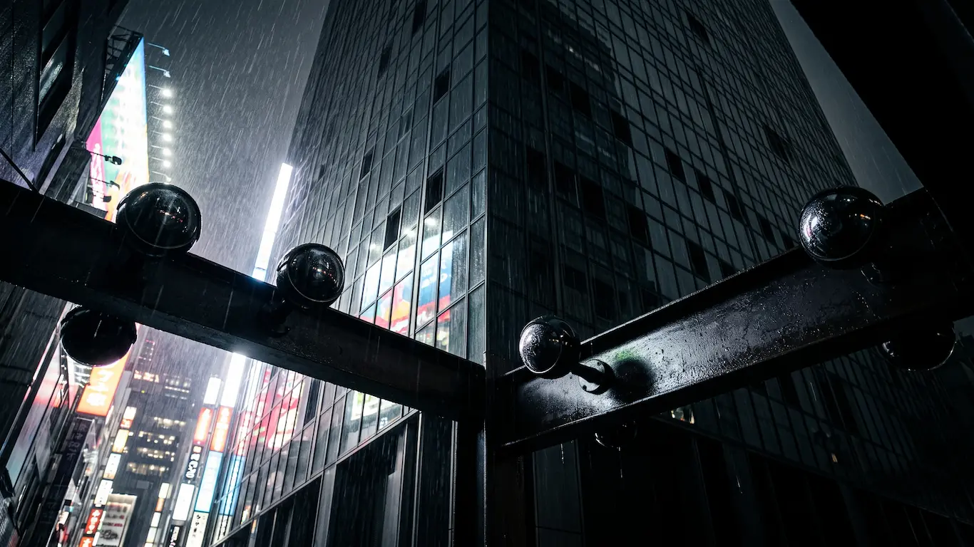 El verdadero origen del cyberpunk anime en japón y el impacto de akira y ghost in the shell - psycho pass sistema sibyl vigilancia tokio Cámaras de seguridad esféricas instaladas en una estructura metálica bajo la lluvia nocturna con rascacielos iluminados de tokio al fondo.